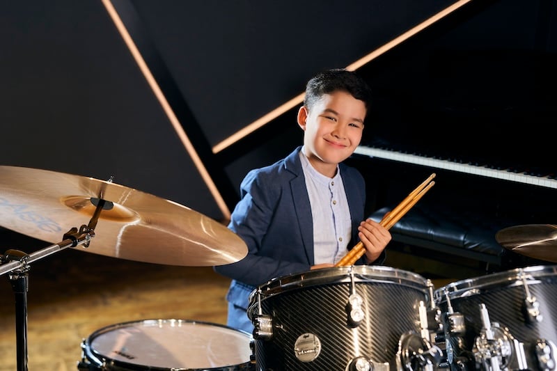 A young student seated behind a drumset is holding two drum sticks in his hands as he confidently smiles.