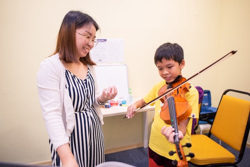 Violin lesson with teacher, in white and young student, in yellow, holding a violin.